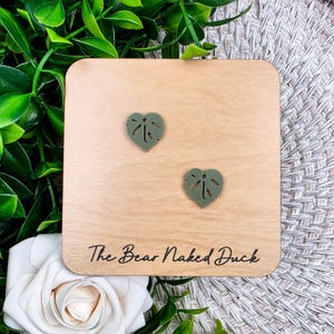 May include: Two olive green heart-shaped earrings with a leaf design are displayed on a light wooden square. The text "The Bear Naked Duck" is printed on the wood. The background includes greenery and a white rose.