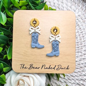 May include: A pair of dangle earrings featuring a yellow sunflower with a brown center, a white bow, and a light blue cowboy boot with a black floral line art design. The earrings are displayed on a square wooden card labeled The Bear Naked Duck.