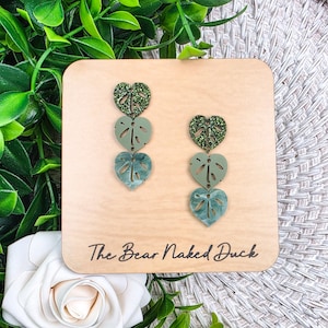 May include: A pair of dangle earrings featuring a layered monstera leaf design. The earrings have a green glitter top, a solid green middle, and a marbled green bottom. The earrings are displayed on a wooden plaque with the text "The Bear Naked Duck".