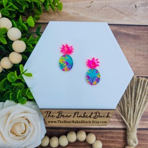 May include: A pair of pineapple earrings with a bright pink and blue tropical print. The earrings are on a white hexagonal display stand with the text "The Bear Naked Duck" and "www.TheBearNakedDuck.Etsy.com".