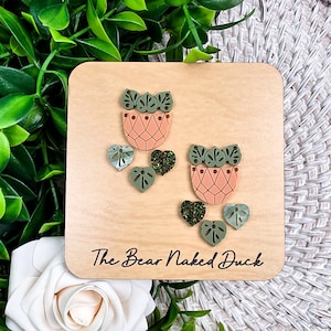 May include: A wooden square with two pink and green plant pot designs and several leaf-shaped and heart-shaped embellishments. The text "The Bear Naked Duck" is printed on the wood. The background includes greenery and a white rose.