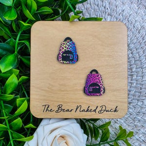 May include: Two colorful backpack-shaped earrings with a leopard print design are displayed on a wooden square. The earrings are in shades of pink, purple, blue, and black. The wooden square has the text "The Bear Naked Duck" engraved on it.