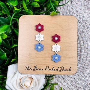 May include: Earrings with a floral design in red, white, and blue, each with a pearl center. The earrings are displayed on a wooden square with the text "The Bear Naked Duck". The background includes greenery and a white rose.