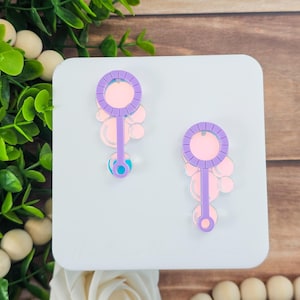 May include: Two bubble wand earrings. Each earring features a purple wand with a circular top and a cluster of pink and clear bubbles. The wands have a light pink center and a blue and teal accent at the bottom.