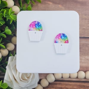May include: A pair of white, snow cone-shaped earrings. The top of each earring is a colorful, rainbow-colored design. The earrings are displayed on a white square with the text "The Snow Cone Co." at the bottom.