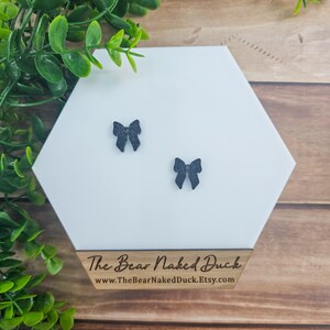 May include: A pair of black bow earrings on a white hexagonal surface. The surface has the text "The Bear Naked Duck" and a website address.