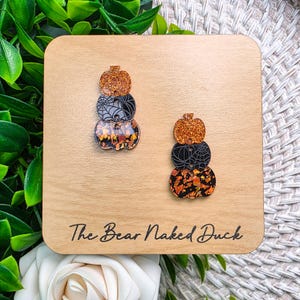 May include: A pair of dangling earrings featuring a stacked pumpkin design. Each earring consists of three pumpkin shapes: a top orange glitter pumpkin, a middle black pumpkin with a spiderweb pattern, and a bottom pumpkin with orange and black flakes.