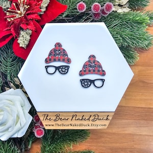 May include: Two wooden ornaments shaped like a hat and glasses. The hat is red and black plaid with a snowflake design. The glasses are black. The ornaments are on a white hexagonal surface with a wooden base that reads "The Bear Naked Duck".