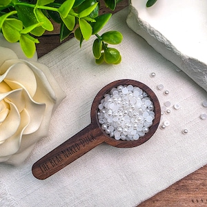 May include: A dark brown wooden scoop with a round bowl filled with small white pearl-like beads. The handle of the scoop is engraved with the text THE BEAR NAKED DUCK in a black sans-serif font. Several loose white beads are scattered nearby.