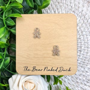 May include: A pair of glittery gingerbread man earrings on a wooden display. The earrings are light brown with iridescent glitter. The wooden display has the text "The Bear Naked Duck" in a cursive font.
