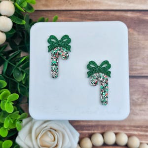 May include: Two Christmas-themed candy cane earrings with green glitter bows. The candy canes are a mix of red, green, and silver glitter. The earrings are displayed on a white surface with a wooden background.