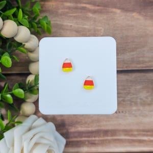 May include: Two candy corn stud earrings. Each earring features a white, orange, and yellow design, mimicking the classic Halloween treat. The earrings are displayed on a white square against a wooden background.