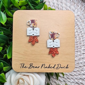 May include: A pair of dangle earrings featuring a floral patterned coffee mug, an open book with black line details, and a glittery orange maple leaf. The earrings are displayed on a light wood square card with the text The Bear Naked Duck.
