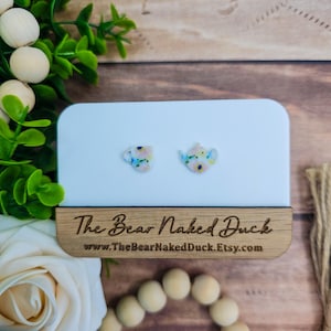 May include: A pair of small, white stud earrings with a floral pattern. One earring is shaped like a teapot, the other like a teacup. The earrings are displayed on a white card with the text "The Bear Naked Duck" and a website address.