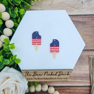 May include: A pair of popsicle-shaped earrings with a patriotic design. The top portion features a blue background with white stars, while the bottom has red and white stripes. The earrings are on a white hexagonal surface, with the text "The Bear Naked Duck" visible.