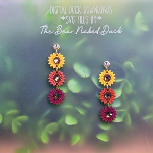 May include: A pair of drop earrings featuring three stacked sunflower-shaped charms in yellow, orange, and dark pink, each centered with a brown crystal. The text DIGITAL DUCK DOWNLOADS *SVG FILES BY* The Bear Naked Duck is visible above.