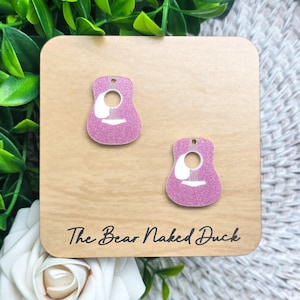 May include: Two pink glittery guitar-shaped charms with white printed details including a sound hole and bridge. The charms are attached to a light brown square display card featuring the text The Bear Naked Duck in black script font.