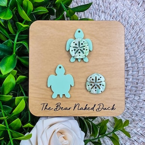May include: Wooden square with three sea-themed acrylic cutouts: two turtles and a sand dollar. The turtles are light blue, one with a patterned shell. The sand dollar has a similar pattern. The text "The Bear Naked Duck" is printed on the wood.