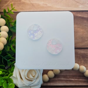 May include: Two circular, iridescent acrylic earring blanks featuring a white snowflake pattern. Each piece has a small circular hole at the top for attachment. The items are displayed on a white square card with the text The Bear Naked Duck.