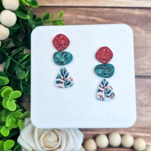 May include: A pair of dangle earrings with a three-part design. The top is a red glittery diamond shape, followed by a teal oval, and a white triangle with a floral pattern. The earrings are displayed on a white square.