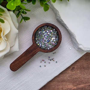 May include: A dark brown wooden scoop filled with small, iridescent, clear rhinestones that reflect multiple colors. Several loose, individual rhinestones are scattered on the white fabric surface near the scoop's handle.