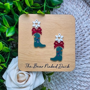 May include: A pair of dangle earrings featuring a white snowflake with a heart cutout, a red glitter bow, and a dark teal cowboy boot adorned with small multicolored rhinestones. The text The Bear Naked Duck is printed on the wooden display card.