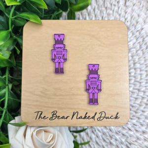 May include: A pair of purple nutcracker earrings on a wooden display. The earrings are shaped like toy soldiers with a hat, mustache, and uniform. The display has the text "The Bear Naked Duck" in a cursive font.