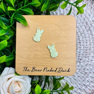 May include: Two light green bunny-shaped earrings with glitter accents are displayed on a wooden square. The earrings are on a wooden square with the text "The Bear Naked Duck" in a handwritten font. The background includes green leaves and a white rose.