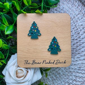 May include: A pair of dark green Christmas tree earrings featuring small, colorful embedded rhinestones. The earrings are displayed on a square light-colored wooden card with the text The Bear Naked Duck printed in black at the bottom.