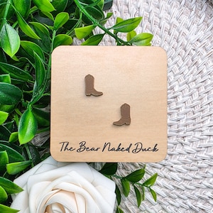 May include: A pair of brown cowboy boot stud earrings attached to a square light wood display card. The card features the text The Bear Naked Duck printed in black script font at the bottom center of the wooden surface.