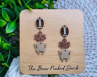 May include: A pair of dangle earrings featuring three stacked charms: a brown and white football, a glittery copper maple leaf, and a beige roasted turkey silhouette. The earrings are displayed on a light wood square card with the text The Bear Naked Duck.