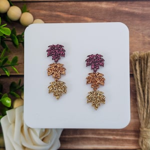 May include: Glittery leaf earrings in shades of purple, copper, and gold. Each earring features three layered leaves, with the top leaf in a deep purple, the middle in copper, and the bottom in gold. The earrings are displayed on a white surface.
