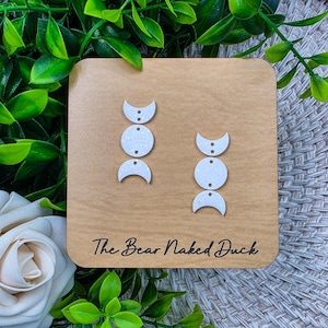 May include: White moon phase earrings with glitter accents are displayed on a wooden square. The earrings are arranged in a vertical line, with a crescent moon shape at the top and bottom, and a circle in the middle. The wooden square has the text "The Bear Naked Duck".