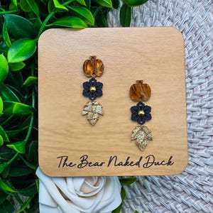 May include: A pair of dangle earrings displayed on a light wood card labeled The Bear Naked Duck. Each earring features a vertical arrangement of a brown marbled pumpkin, a black flower with a gold center, and a textured gold leaf charm.