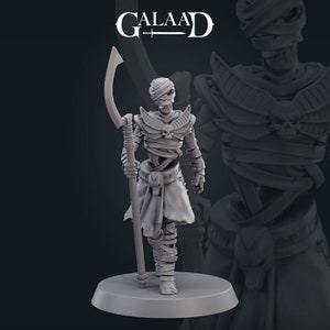 May include: A gray painted miniature figurine of a mummy warrior holding a curved staff. The mummy is wearing a head wrap and a loincloth. The figure is standing on a round base.
