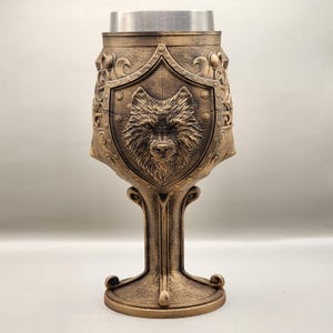 May include: A gold-colored goblet with a wolf's head sculpted on a shield. The goblet has a decorative base and a silver-colored top.