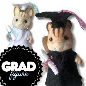 May include: Two graduation-themed animal figurines. One is dressed in a white graduation gown and cap with a blue tassel, holding a scroll. The other is in a black gown and cap with a pink tassel, also holding a scroll. The image includes the text "GRAD figure".