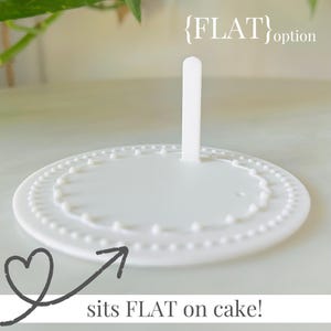 Op de afbeelding: Een witte taarttopper met een platte basis en een verticale steun. De basis heeft een decoratieve, parelrand. De tekst "{FLAT} option" staat bovenop de topper, en "sits FLAT on cake!" staat eronder.