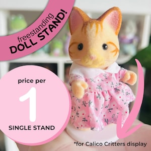 May include: A Calico Critters cat figure in a pink floral dress, on a white base. The image includes text: "freestanding DOLL STAND!" and "price per 1 SINGLE STAND". For Calico Critters display.