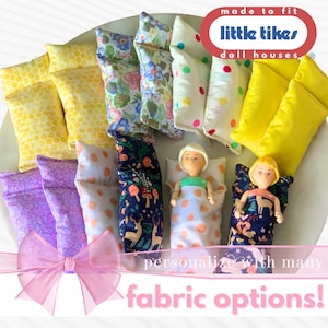 May include: A collection of miniature fabric sleeping bags and pillows for dollhouses, featuring various patterns including yellow honeycomb, floral, polka dot, solid yellow, purple floral, and woodland prints with two small dolls tucked inside.