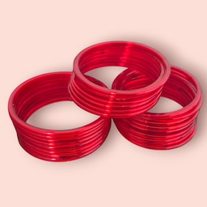 May include: Three stacks of bright red bangles, each with a glossy finish, are arranged on a light pink background. The bangles are circular and appear to be made of a solid material.