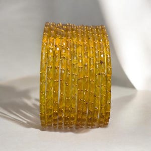 May include: A stack of yellow-gold bangles. Each bangle is made of faceted, translucent beads with small gold-colored accents. The bangles are stacked together, creating a shimmering effect. The background is a soft, neutral color.