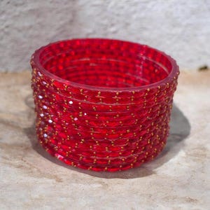 May include: A stack of red glass bangles with gold-colored accents. The bangles are transparent and faceted, catching the light. The jewelry is stacked on a neutral-colored surface.