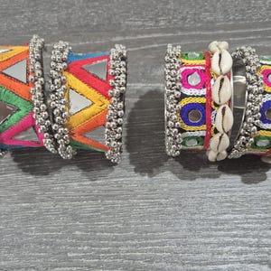 May include: Two pairs of colorful embroidered bracelets. One pair features a rainbow pattern with mirrored accents and silver bead trim. The other pair has a design with shells and mirrored accents, also with silver bead trim.
