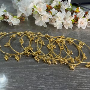 May include: A set of six thin gold-toned metal bangles, each featuring small, dangling spherical charms spaced evenly along the circular band. The bracelets are arranged in an overlapping cluster, showcasing a polished, reflective metallic finish.