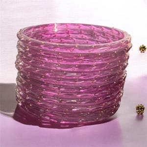 May include: A stack of translucent magenta glass bangles featuring a faceted surface texture and small, evenly spaced gold-colored metallic accents embedded along the outer circumference of each individual circular bracelet.