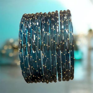 May include: A set of ten translucent dark blue bangles arranged in a vertical stack. Each bangle features a faceted, textured surface with small, evenly spaced gold-colored metallic dots embedded along the ridges of the faceted design.