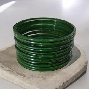 May include: A stack of dark green bangles, resting on a light gray, textured surface. The bangles are circular and appear to be made of a glossy material. The stack is composed of multiple bangles, creating a layered effect.