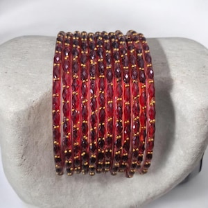 May include: A stack of red beaded bracelets with gold-colored accents. The bracelets are made of small, faceted beads and are arranged on a light gray, textured surface. The bracelets are a deep red color.