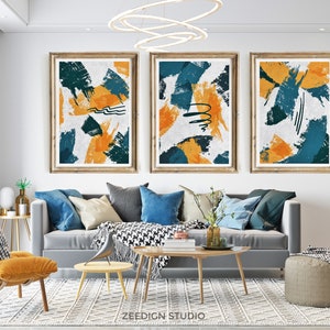 以下が含まれることがあります： 白地にターコイズと黄色の大胆な筆致が施された抽象的なアートプリント3枚。プリントは明るい木製のフレームに収められ、灰色のソファの上の白い壁に掛けられています。